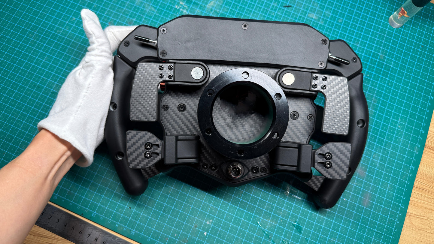 Ferrari 499P endurance sim racing wheel rear view showing carbon fiber backplate, central mounting bore and magnetic paddle shifter mechanism, held in gloved hand.
