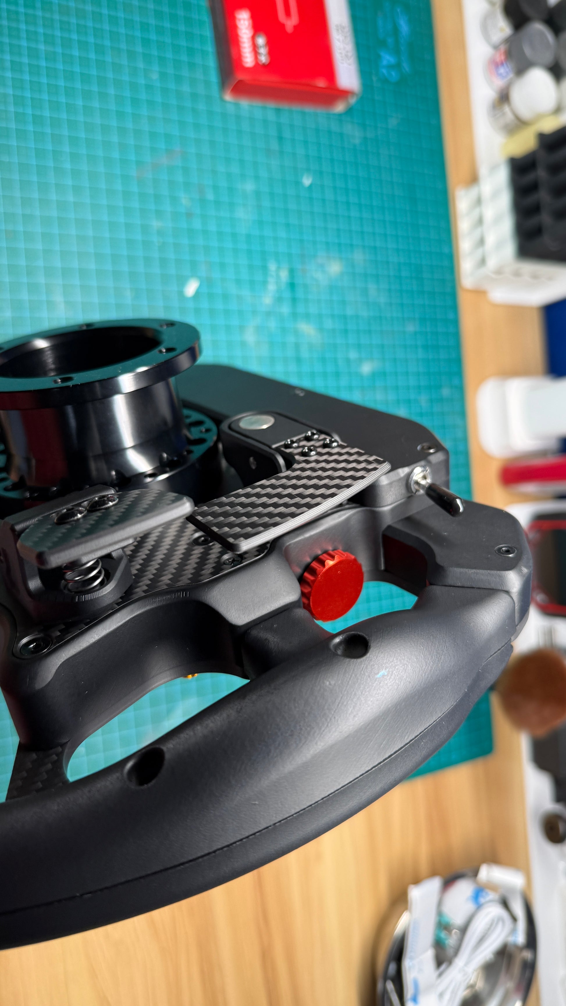 Ferrari 499P endurance sim racing wheel close-up of the carbon fiber magnetic shift paddle and red anodized rotary encoder detail beside the quick-release hub.